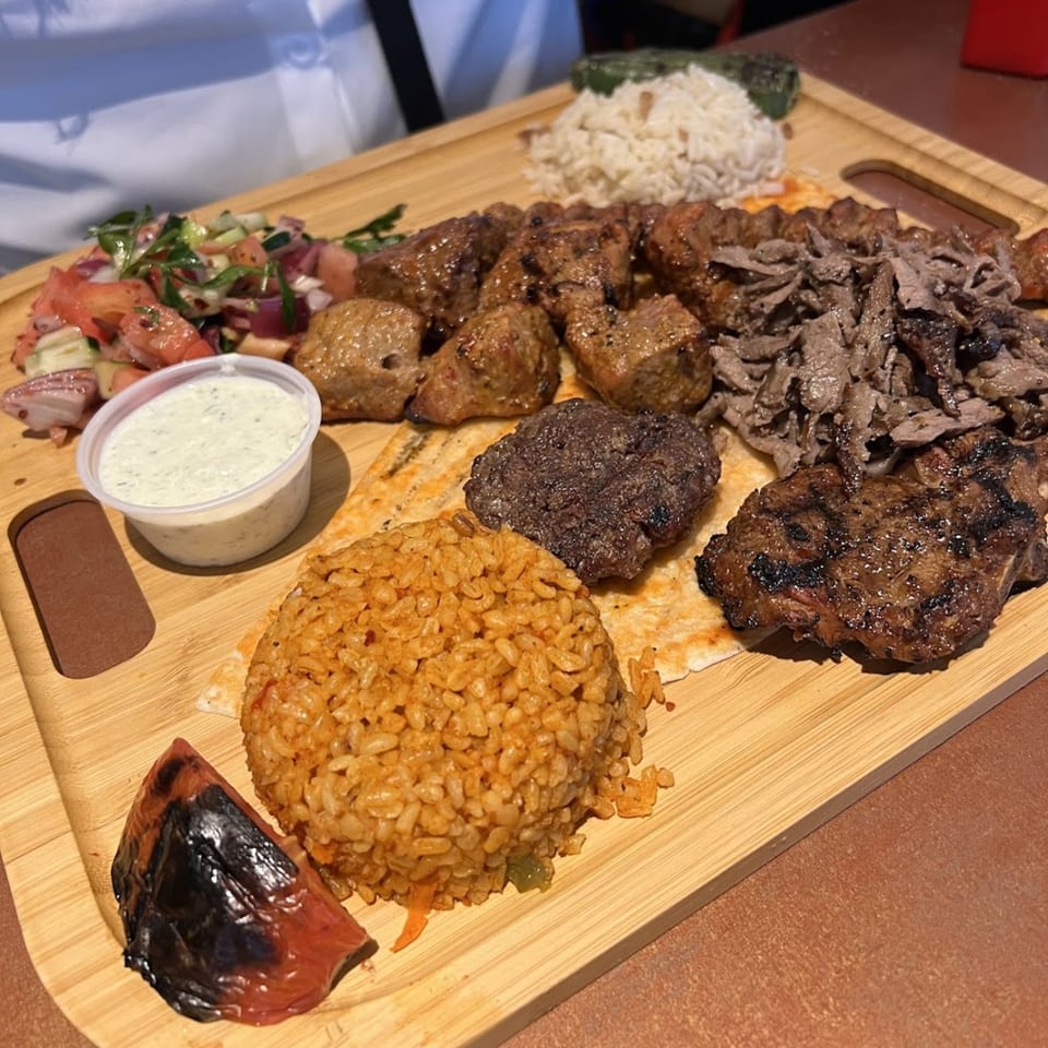 A Turkish mixed grill platter with various kebabs, rice, salad, and sauce on a wooden board.