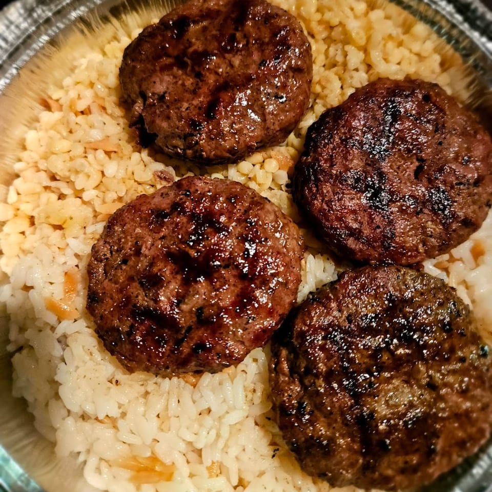 Four grilled Turkish kofte patties with char marks, served over white and bulgur rice in a foil container.