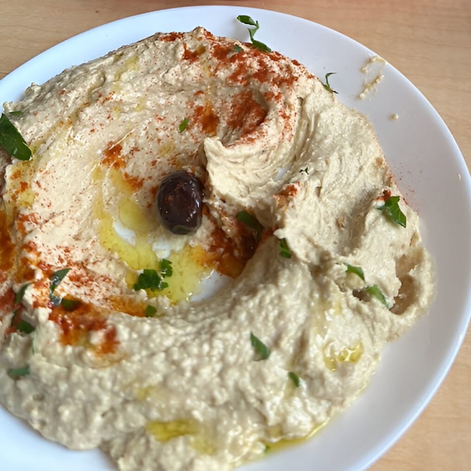 Creamy Turkish hummus with a black olive, paprika, olive oil, and parsley on a white plate.