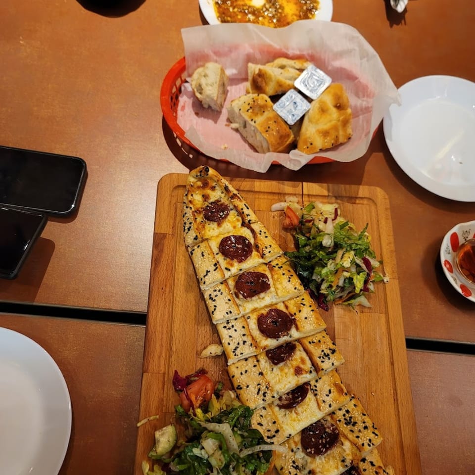 Turkish pide with sucuk and sesame seeds, served with salad, bread, and tea on a wooden table.