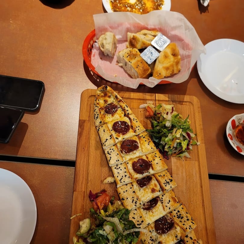 Turkish pide with sucuk and sesame seeds, served with salad, bread, and tea on a wooden table.