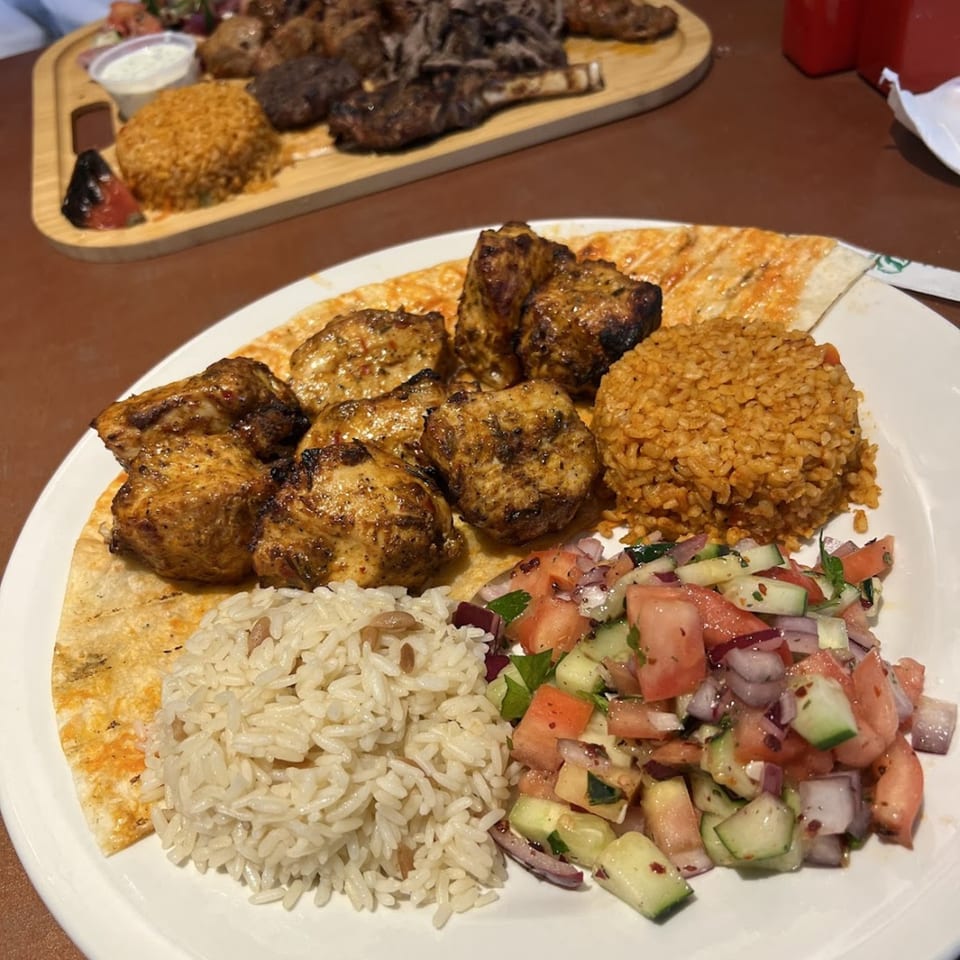 Turkish chicken kebab plate with rice, bulgur, salad, and flatbread, with a meat platter in the background.